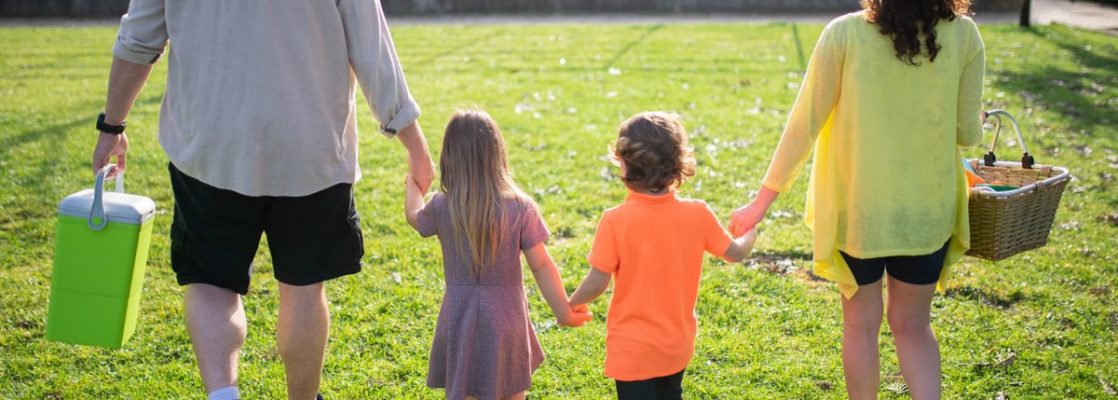 A family of four walking hand in hand through a sunny park carrying picnic supplies.