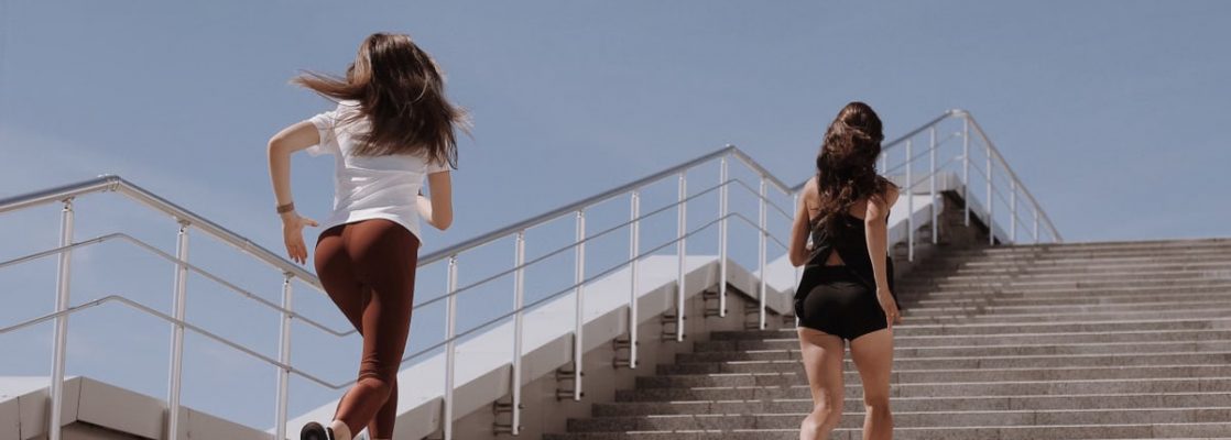 Two women running up outdoor stairs for fitness under a clear blue sky.